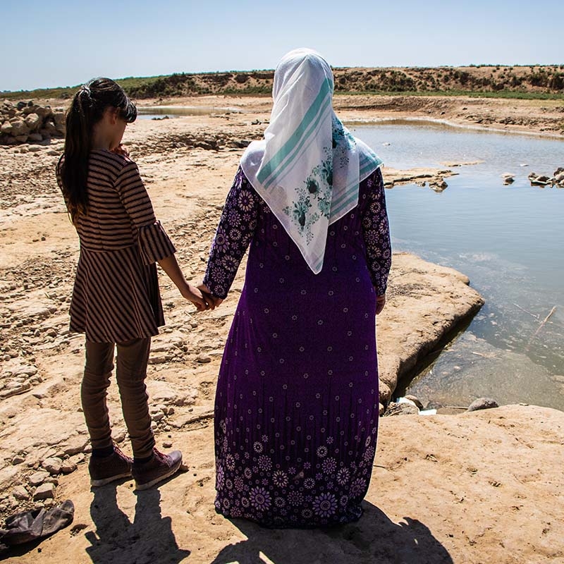 A mother and her daughter hold hands while staring out over a dried and drought-ravaged landscape.