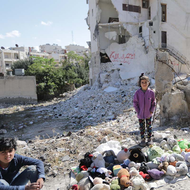 In Syria, a girl stands in front of a building that has been destroyed by airstrikes. 