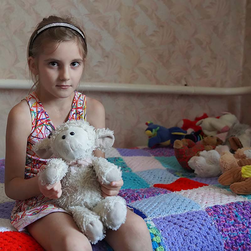 A young girl who fled conflict in Ukraine sits on a bed and holds a stuffed animal. 