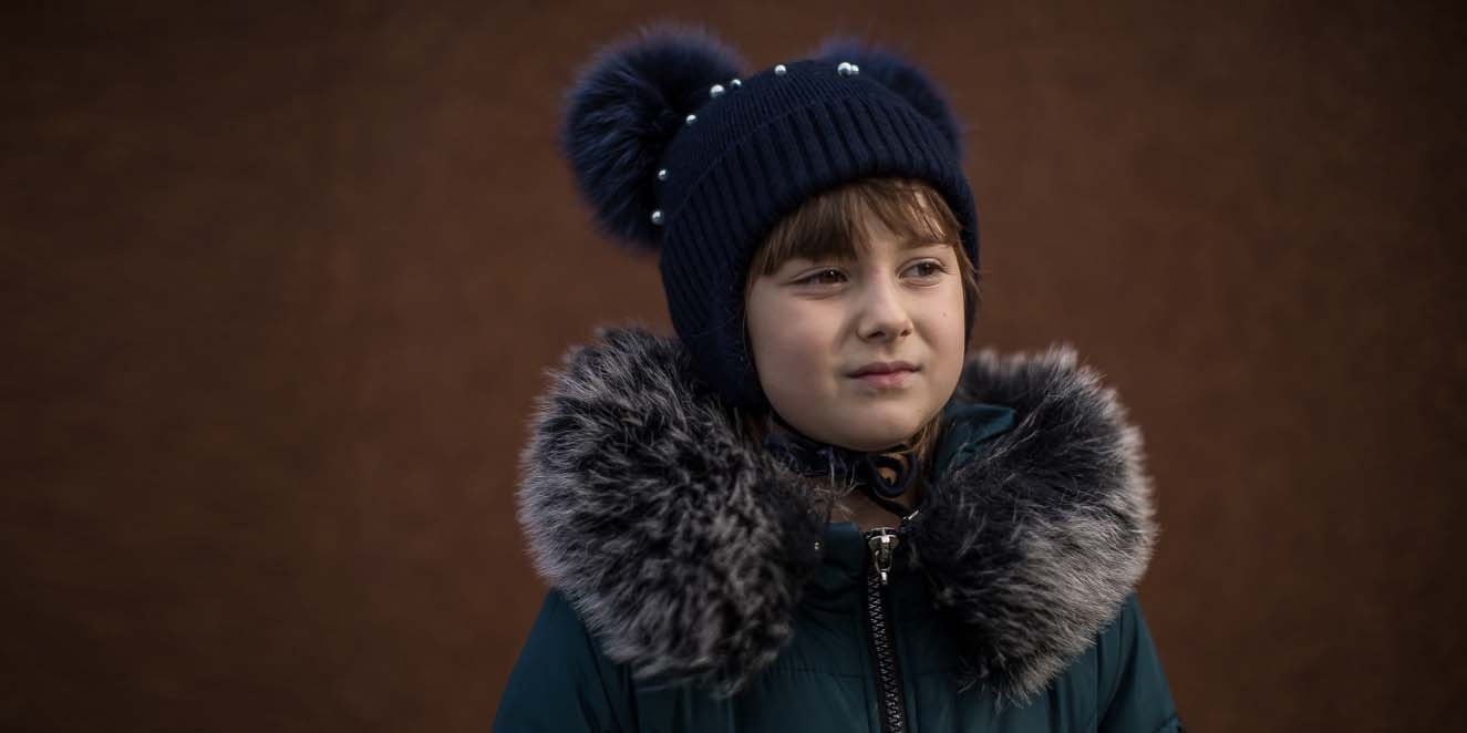 In Ukraine, a girl wears warm winter clothing while standing alone against a dark background.