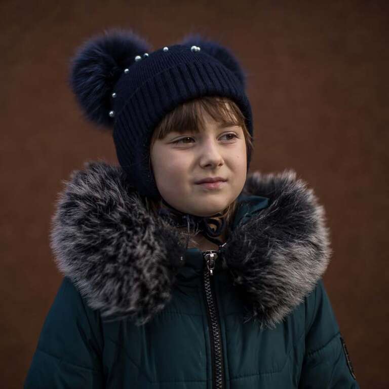 In Ukraine, a girl wears a hat and coat while standing alone. 