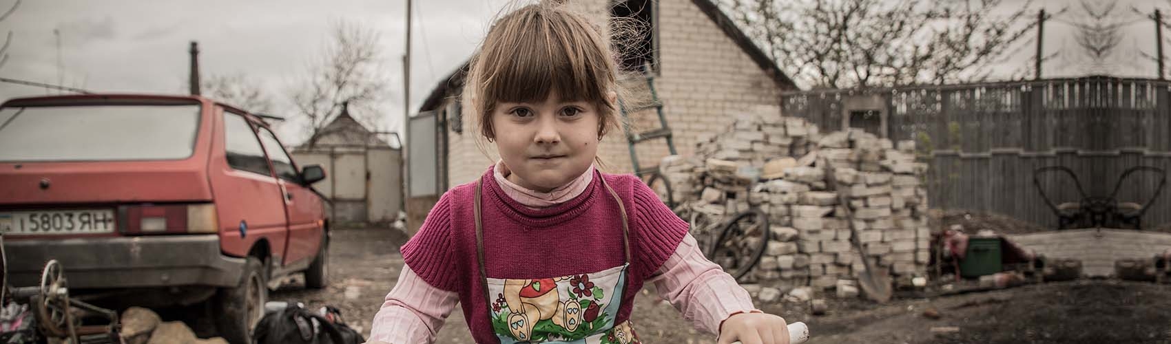 In Ukraine, a girl rides a bike near a crumbled wall and dirt path.