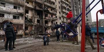 In Ukraine, a girl swings on a swing in a playground near a building that has been demolished by a attack.