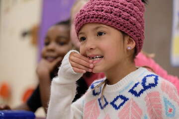 A girl touches her hands to her mouth while listening intently with other students in her classroom.