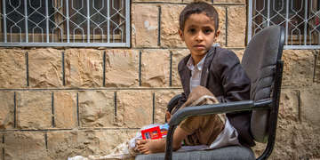 A young boy named Ismail sit on a chair outside a brick building with bars on the windows. The boy’s right leg is wrapped, from his toe to above his knee, in white bandages. The boy was on a school bus, riding through Yemen when his bus was bombed by an aircraft belonging to the SLC (Saudi-Led Coalition). Save the Children’s Stop the War on Children campaign draws attention to children living in conflict and calls on world leaders to take action to protect children living in war zones. 