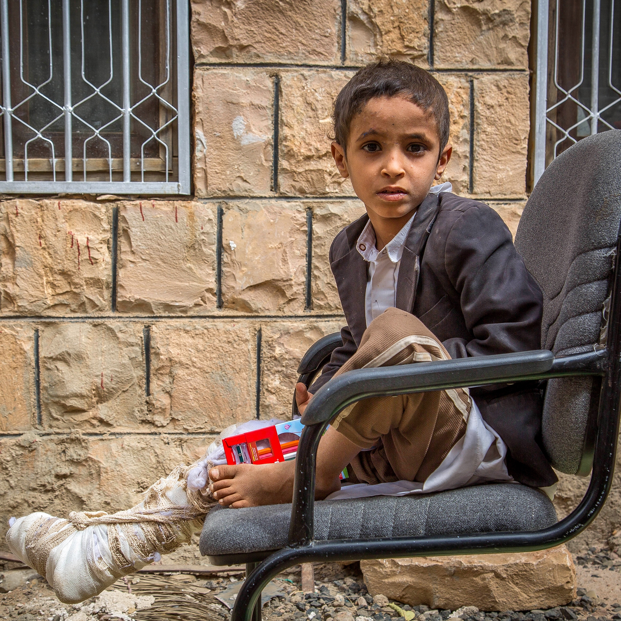A young boy named Ismail sit on a chair outside a brick building with bars on the windows. The boy’s right leg is wrapped, from his toe to above his knee, in white bandages. The boy was on a school bus, riding through Yemen when his bus was bombed by an aircraft belonging to the SLC (Saudi-Led Coalition). Save the Children’s Stop the War on Children campaign draws attention to children living in conflict and calls on world leaders to take action to protect children living in war zones. Photo credit: Mohammed Awadh / Save the Children, Sept 2018. 