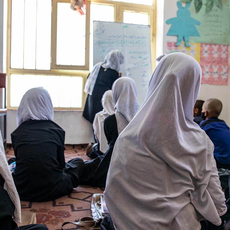 In Afghanistan, female teacher stands at the head of a classroom where boys and girls are seated.