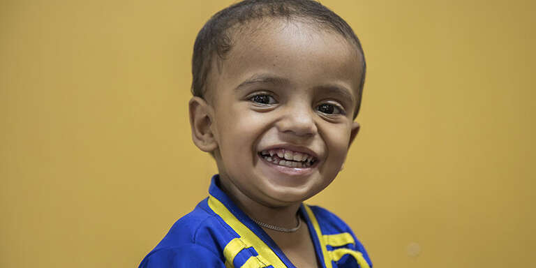 A child in a blue shirt against a yellow background smiles at the camera.