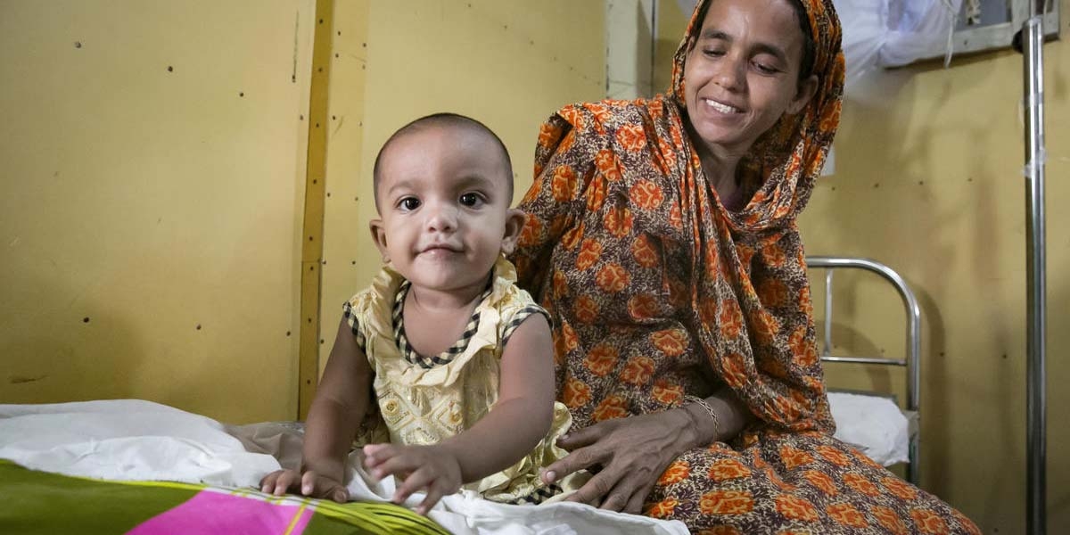 An 8-month old baby smiles while sitting on a hospital bed with his mother in Bangladesh.
