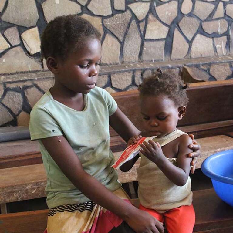 A 9-year old girl sits with her baby sister who eats a food supplement on the floor of a home in DRC.