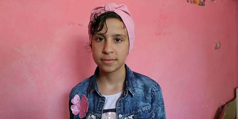 A girls in a blue jacket and pink headband stands in front of a pink wall.