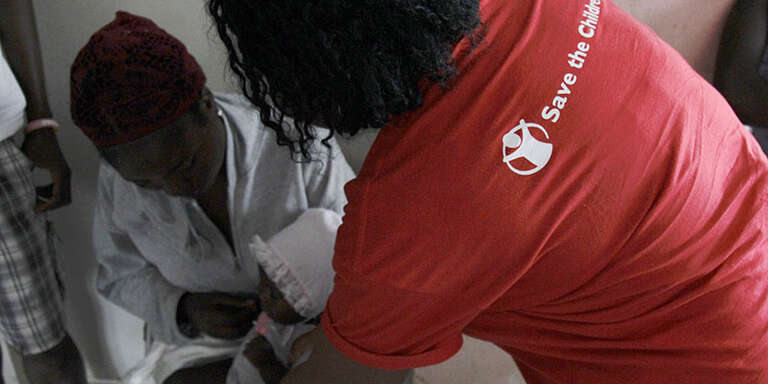 A Save the Children staff member leans over a doctor treating a baby.