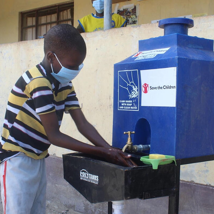 Kenya, a little boy in a striped shirt and face mask, washes his hands at a Save the Children hand washing station