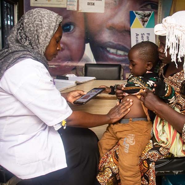 A healthworker holds a a table while conducting an assessment with a child sitting on his mother's lap.