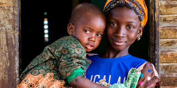  12-year-old Gniré holds her younger brother outside of their family home in Sikasso, Mali. In 2017, Gniré was diagnosed with malaria but survived thanks to treatment. Save the children provided Gniré  with medication a mosquito net and works globably to reduce child deaths related to preventable diseases like malaria. Photo credit: Victoria Zegler | Save the Children, Nov 2016.
