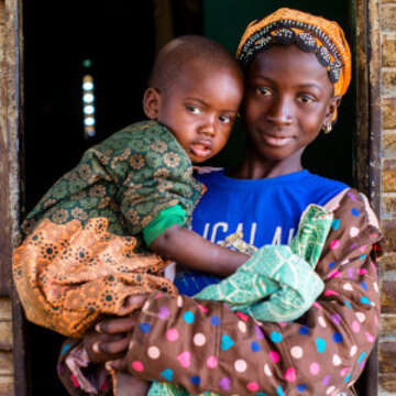12-year-old Gniré holds her younger brother outside of their family home in Sikasso, Mali. In 2017, Gniré was diagnosed with malaria but survived thanks to treatment. Save the children provided Gniré  with medication a mosquito net and works globably to reduce child deaths related to preventable diseases like malaria. Photo credit: Victoria Zegler | Save the Children, Nov 2016.