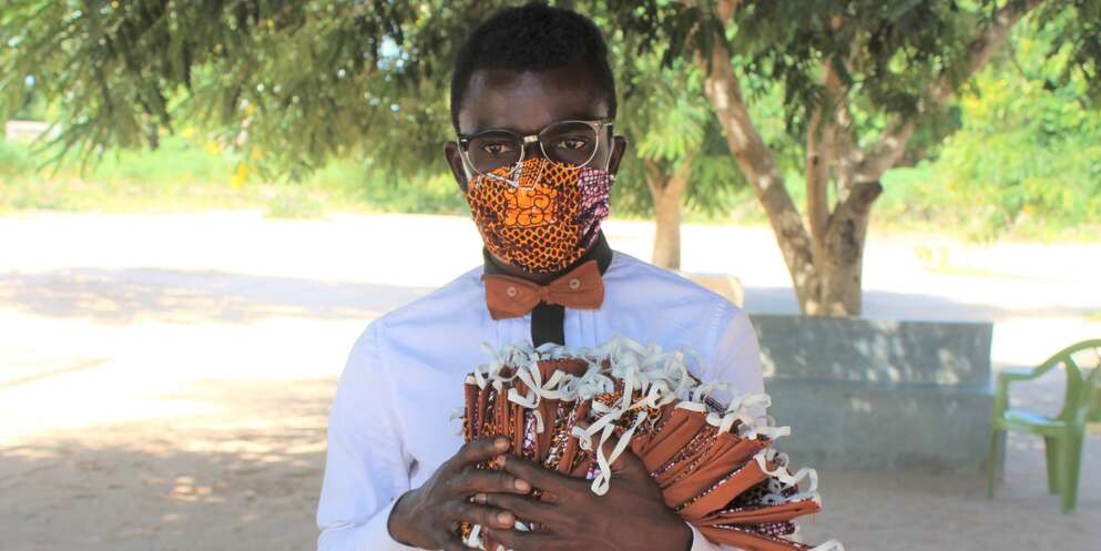 A young man in Mozambique wears a handmade face mask while holding an arm's load of additional masks. 