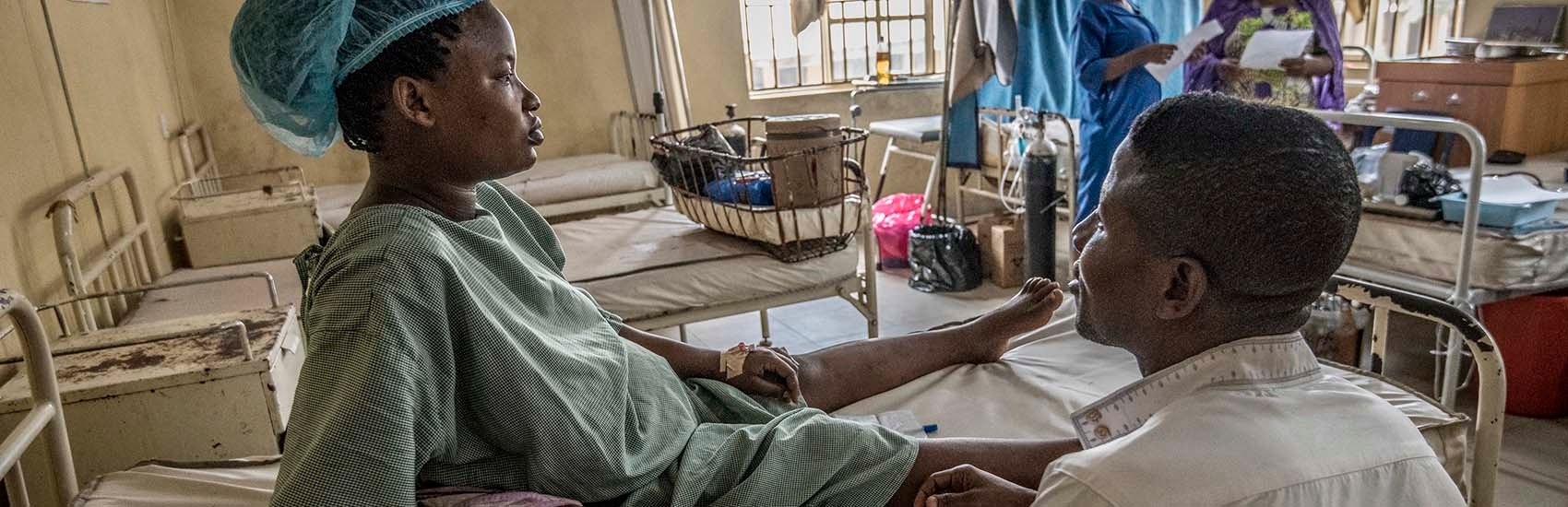 A woman wearing a blue hospital gown sits on a hospital bed in the maternity ward and looks straight ahead while a doctor sits beside her. Other female patients at the Okene Zonal Hospital in Nigeria can be seen in the background along with several white hospital beds. Many of the doctors at this hospital are on strike. The maternity ward use to be very busy but with the doctors on strike this state hospital is practically empty. Photo credit: Save the Children 2020.