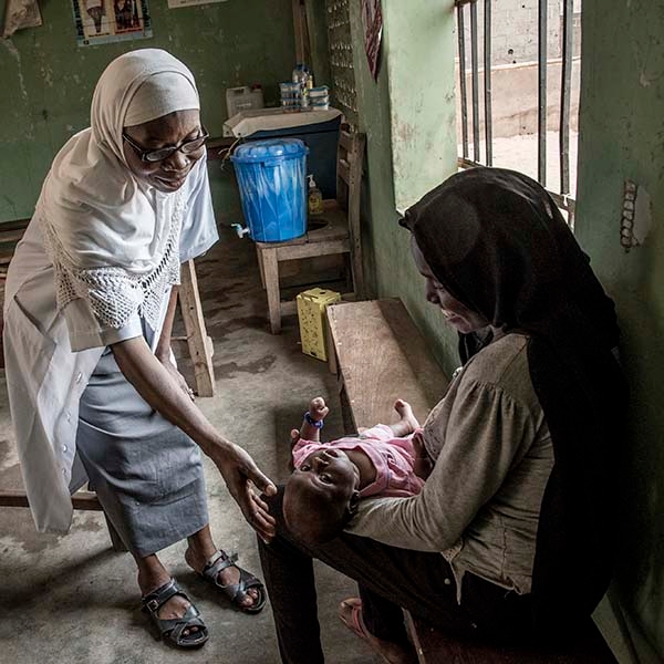 A mother sits with her 3-month old baby at Sarkin Noma Primary Health Center (PHC) in Nigeria. The 19-year old new mom is getting advise from the head community health officer. The health officer is placing her hand near the baby’s head and smiling. The new mom is smiling as well. 