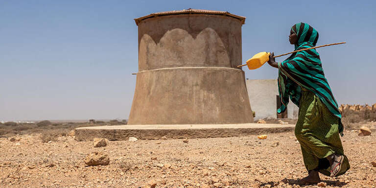 A teenage girl walks on dry land past a concrete well carrying a yellow water jug.