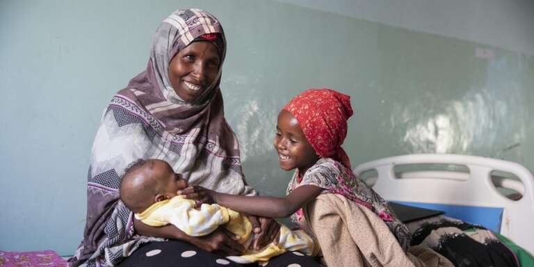 A woman smiles and holds her baby as an older child stands nearby. 