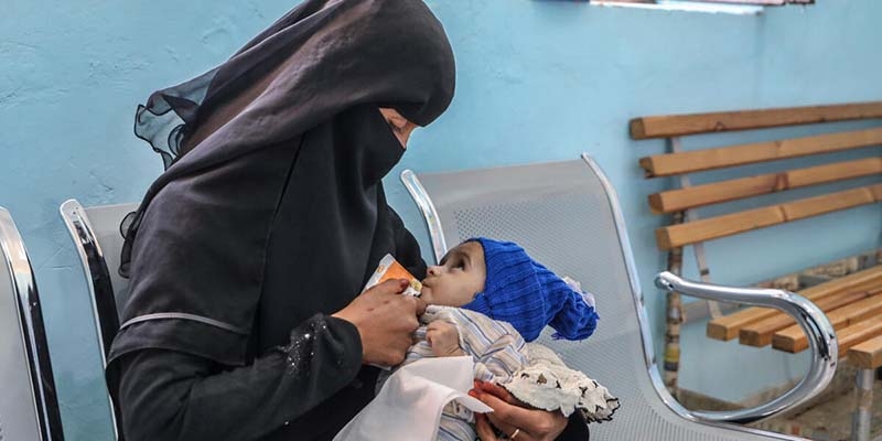 A woman in a black head and face covering holds her child and feeds her baby, who is wearing a blue hat.