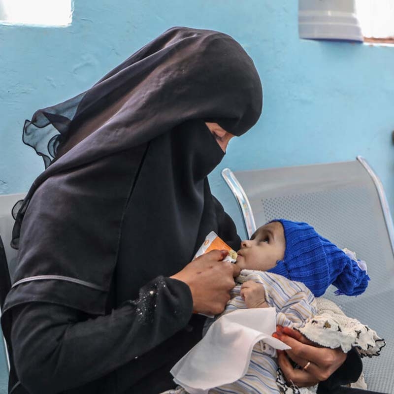 Yemen, a caregiver feeds a baby in a blue hat