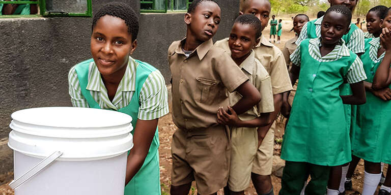 A girl leans over to wash her hands out of a white bucket and looks at the camera. A line of girls stand behind her.