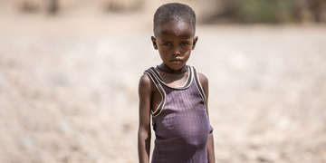 A four-year old boy stands in a dry field in northern Kenya and wears a purple shirt, covered in dust. The boy was suffering from acute malnutrition and identified by a Save the Children outreach team. He was registered for treatment and given a supply of ready-to-use therapeutic food packets. In the months since he began treatment, the boy’s condition has improved greatly. Photo credit: Colin Crowley / Save the Children, June 2017. 