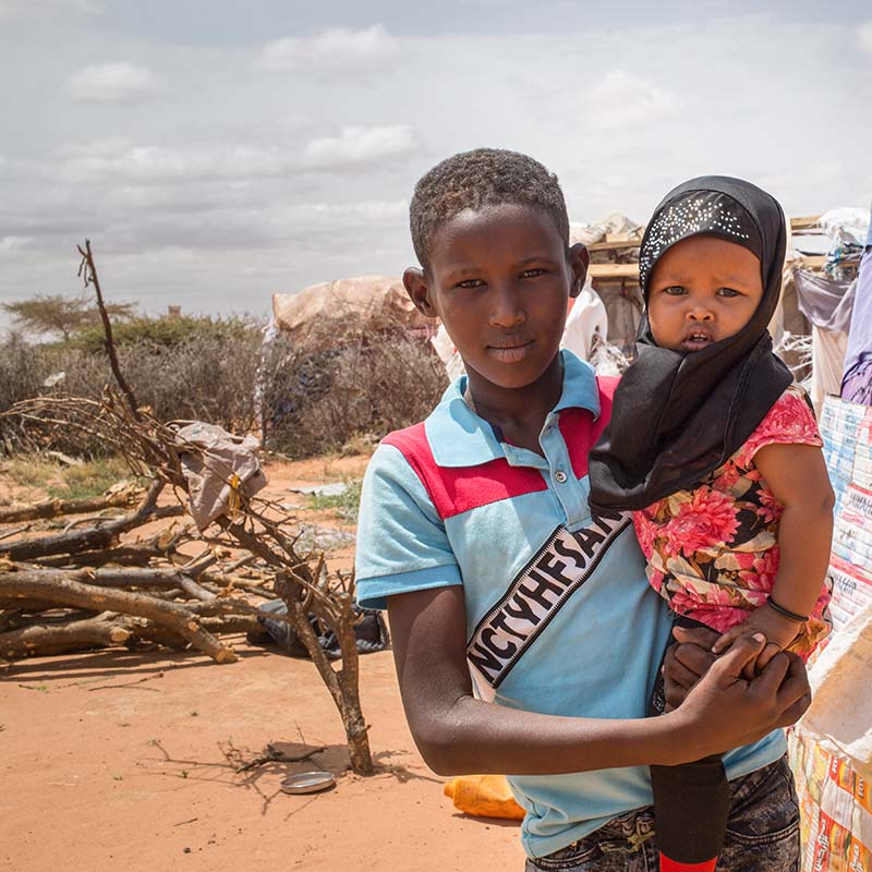 In Somalia, a pair of siblings stand together in front of a dry landscape where a shelter is in place. 