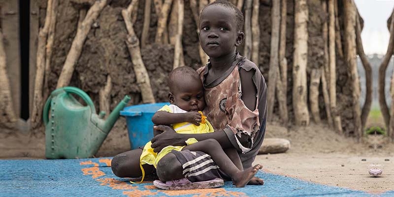In South Sudan, a child holds a baby while sitting outside on a blue mat. 