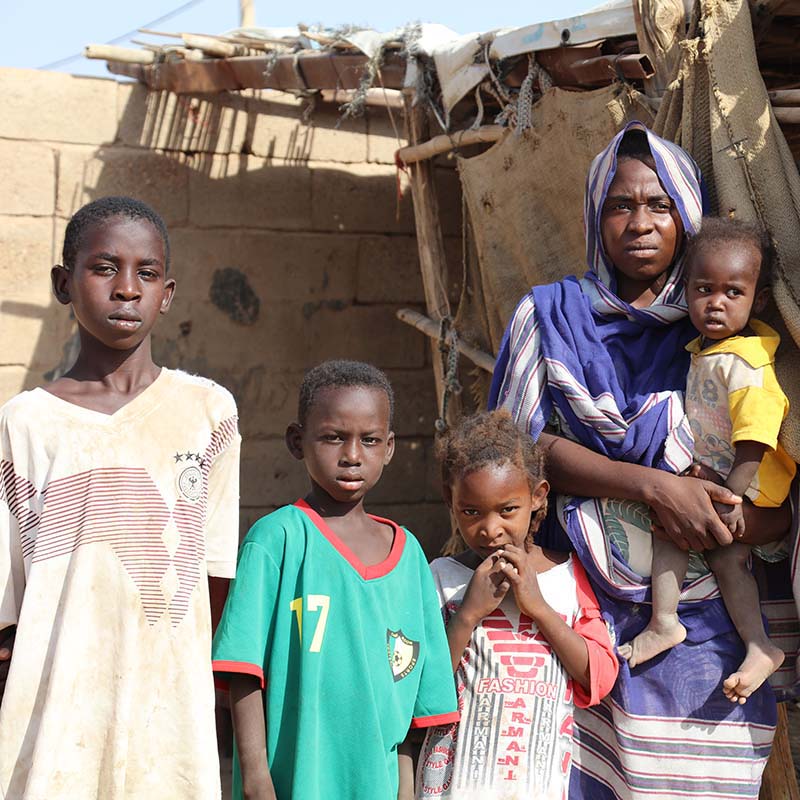 In Sudan, a family stands in front of their home as a dry landscape can be seen in the background.