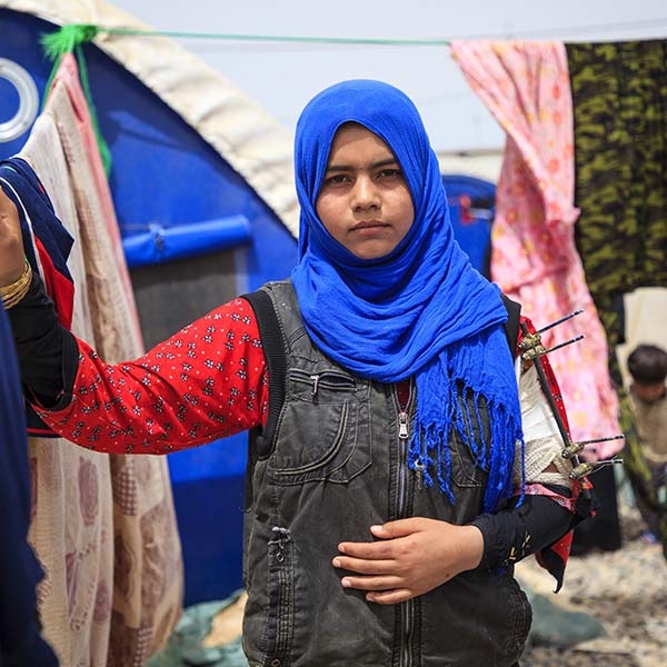 Fourteen-year-old Sulaima stands outside her family’s refugee tent, wearing a blue head scarf and red, flowered shirt – her arm in a cast, with pins. 