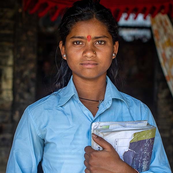 Nepal, a young girl in a blue shirt holding a book looks into the camera
