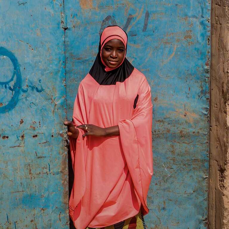 In Niger, a young girl stands against a brown cement wall on a bright day.