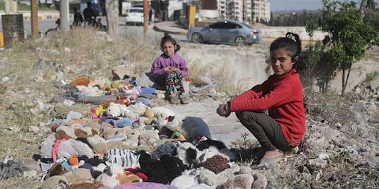 Two girls sit among rubble selling toys to earn money for their family after their home in Syria was bombed
