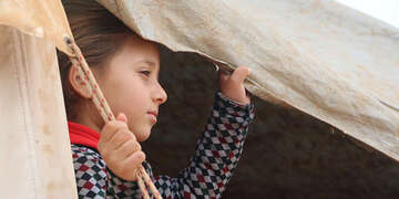 A young girl looks out of a tent while holding the canvas with her hand. 