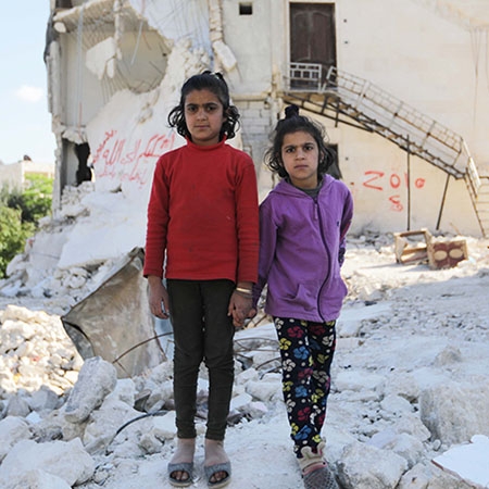 In Syria, two young girls stand atop a pile of ruble and debris caused by deadly conflict. 