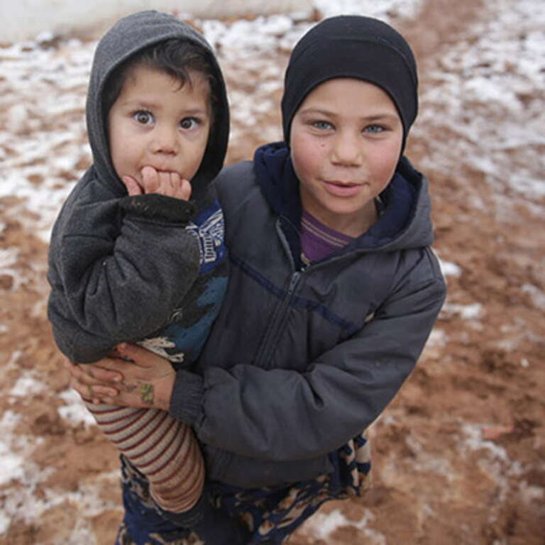 A young child is held by an older child while standing in a refugee camp in Syria.