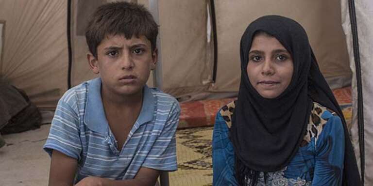Two young siblings sit together in a tent in a refugee camp in Syria.