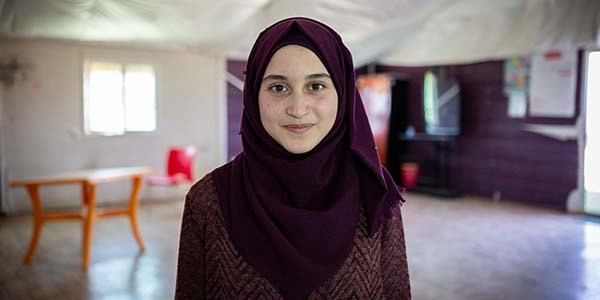 A 14-year old girl stands in a tent with a table and smiles. The girl is from Syria and her family fled to a refugee camp in Za’atari where she now attends school.