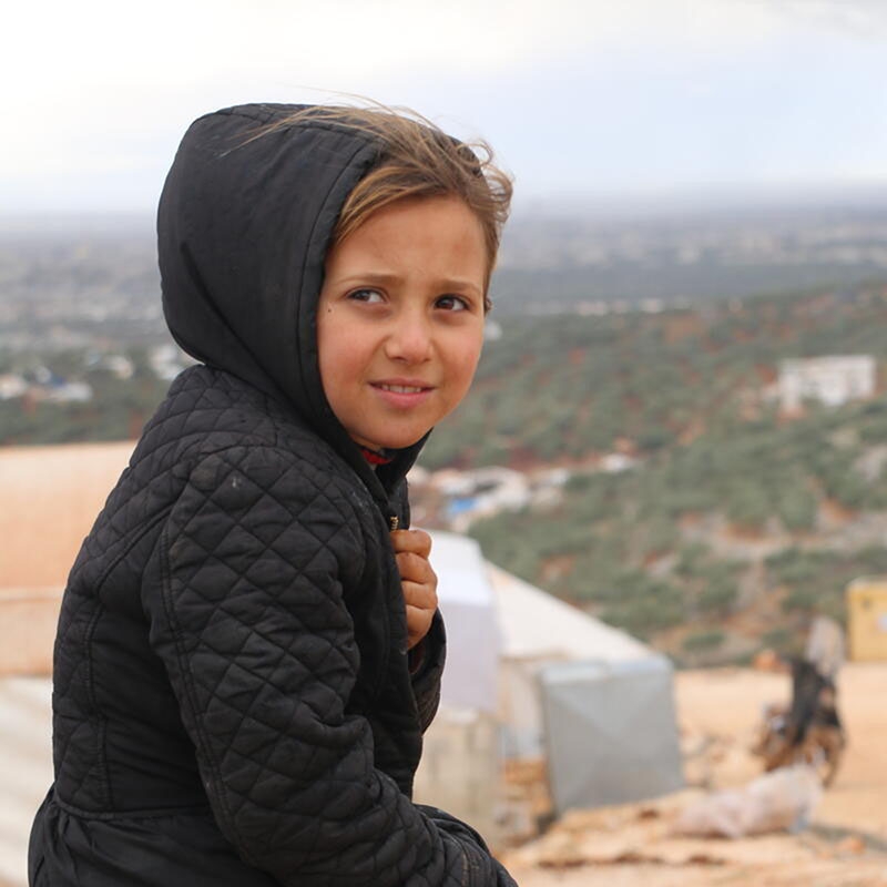 Syria, a little girl in a black winter coat stands outside her tent