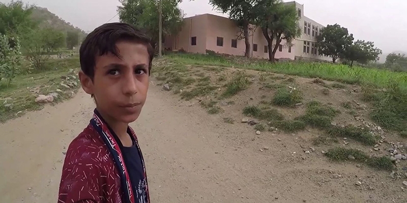 A Yemeni boy in a red and black shirt looks at the camera, and his school, a large beige building is in the background. 