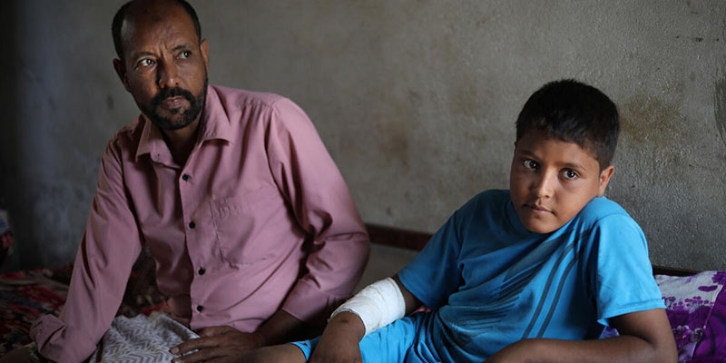 A teenage boy in a blue shirt and arm bandage leans on a bed. His father, in a pink shirt, sits next to him.