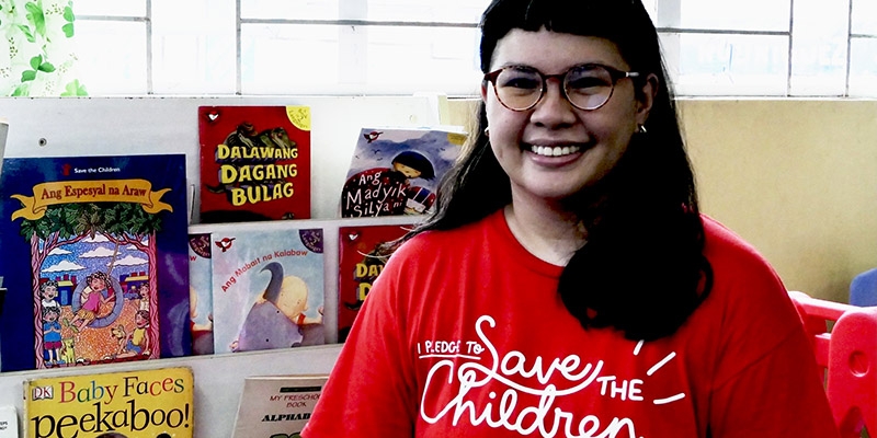 Save the Children worker smiles at the camera in front of a shelf filled with children's books.