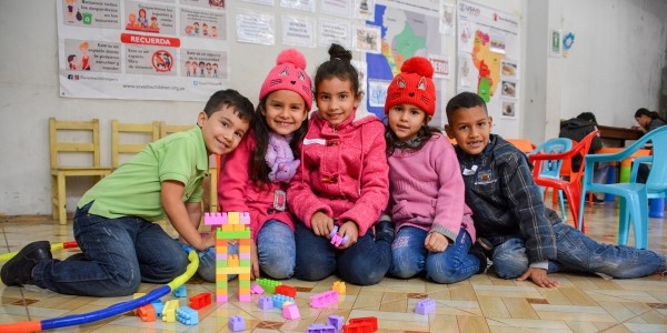 A smiling group of children gather together in an area decorated with educational posters, building blocks and bright furniture.