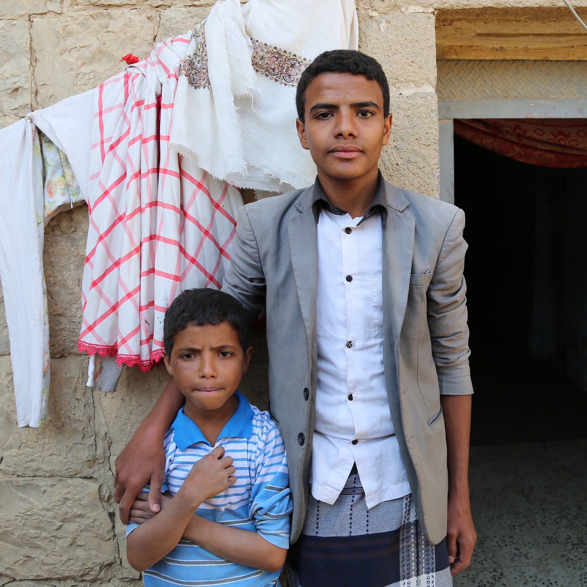 Hamza*, 16, with his brother Hani*, 12, outside their home in Amran, Yemen. Hamza*, helps provide for his mother and five siblings. He was injured on the first day of airstrikes in Sana'a when a shell injured him and broke his leg. Photo Credit: Mohammed Awadh/Save the Children.