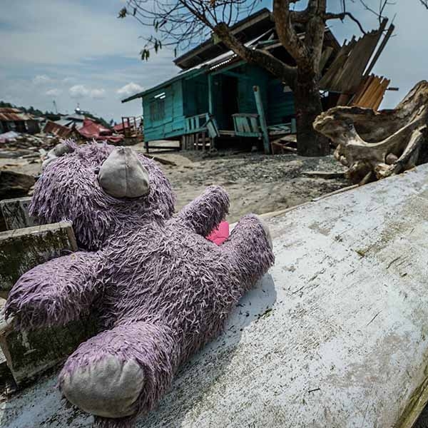 A teddy bear lays strewn across wreckage on the beach after the Sulawesi Indonesia earthquake and tsunami. In the background, a family's home is in shambles, torn apart by the tsunami's crushing wall of water. Photo credit: Thomas Gustafian / Save the Children 
