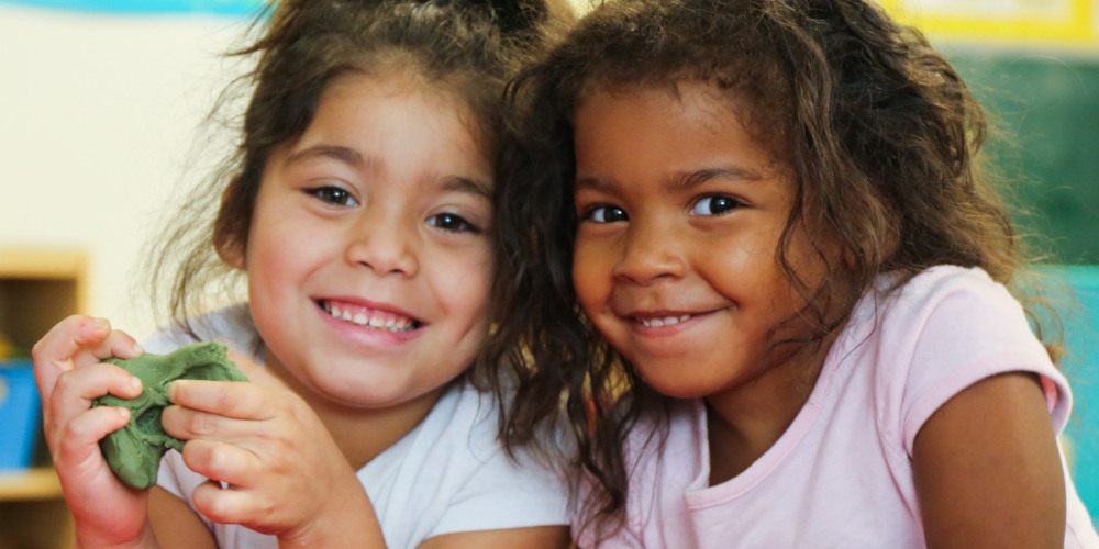 A pair of friends play and smile at a table in a classroom. 