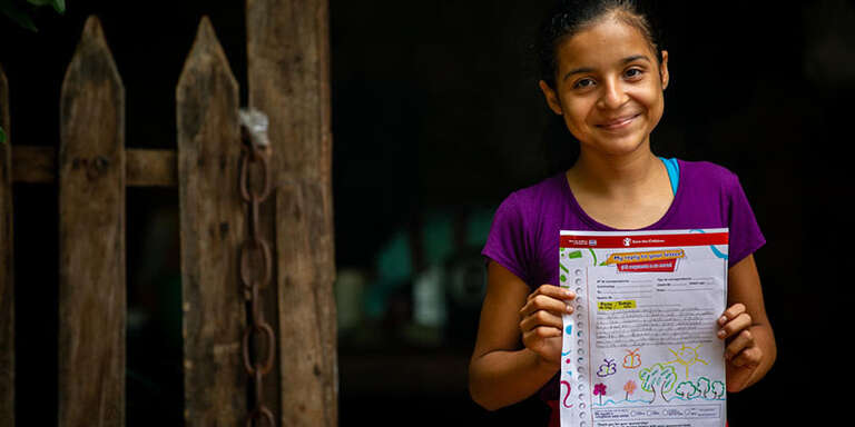 A girl in purple shirt standing next to a wood m fence holds up a letter written to her sponsor and smiles.